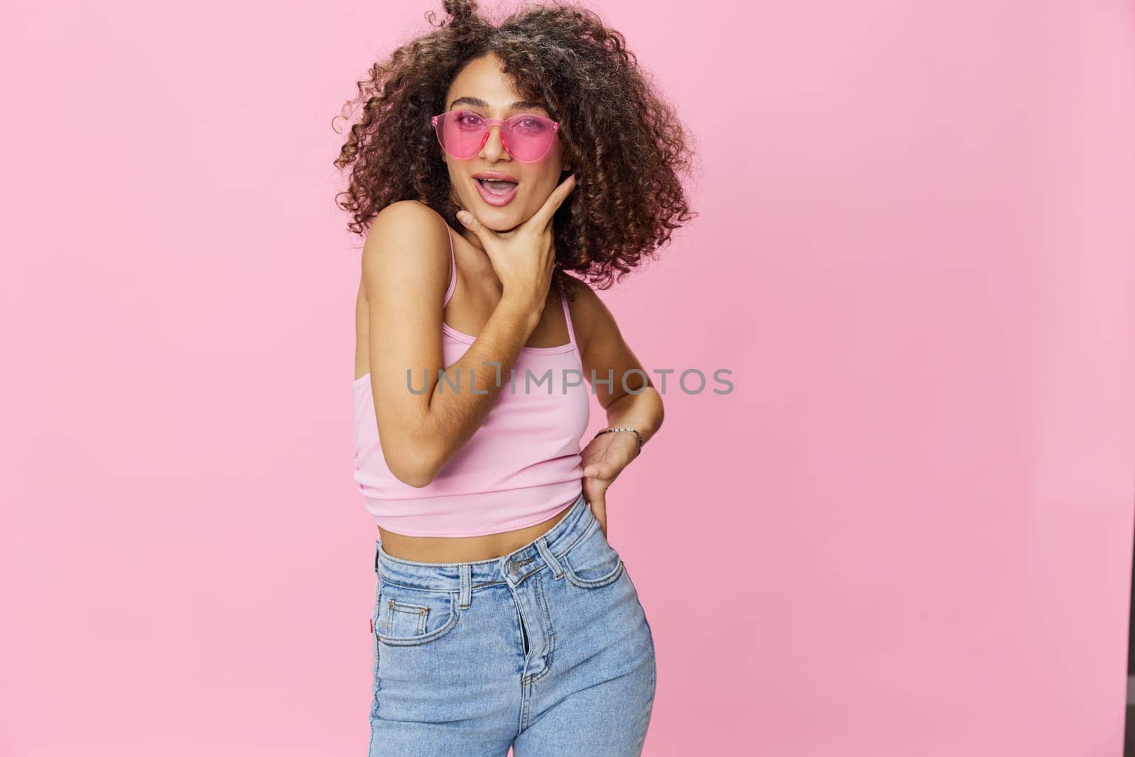 Happy woman with curly hair in a pink T-shirt and jeans on a pink background with sunglasses with a beautiful tan, DJ party, copy space. High quality photo