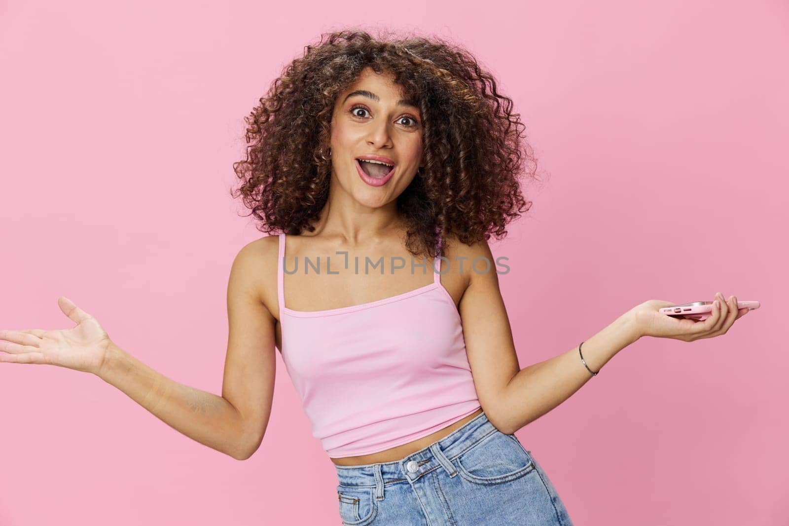 Woman blogger holding phone with curly hair in pink top and jeans poses on pink background, copy space, technology and social media. High quality photo
