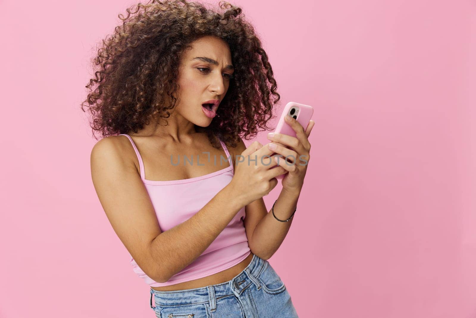 Woman blogger holding phone with curly hair in pink top and jeans poses on pink background, copy space, technology and social media. High quality photo