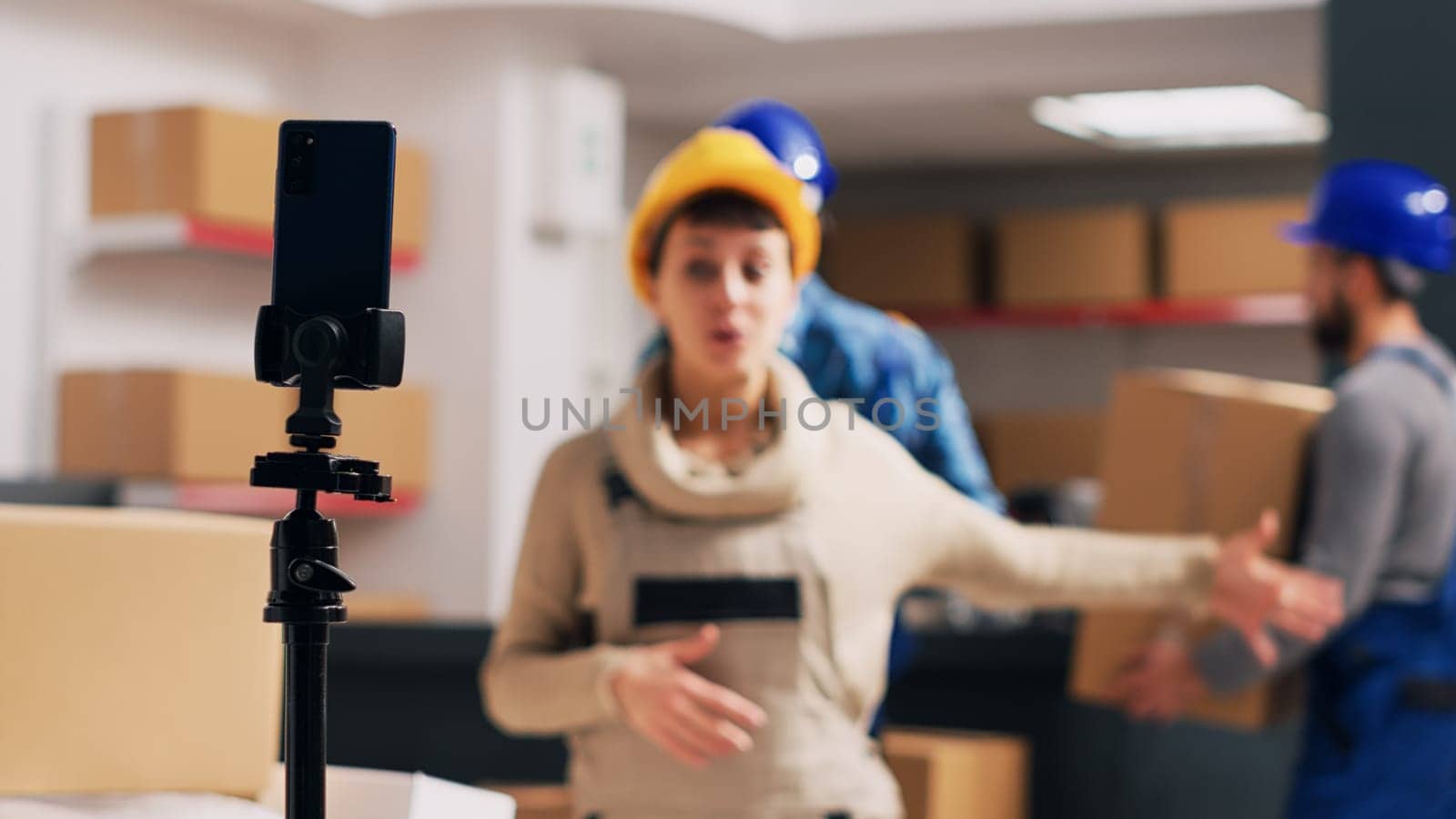 Female employee filming video ad in storage room, advertising depot products on smartphone app. Young woman in overalls recording online social media video, industrial space.