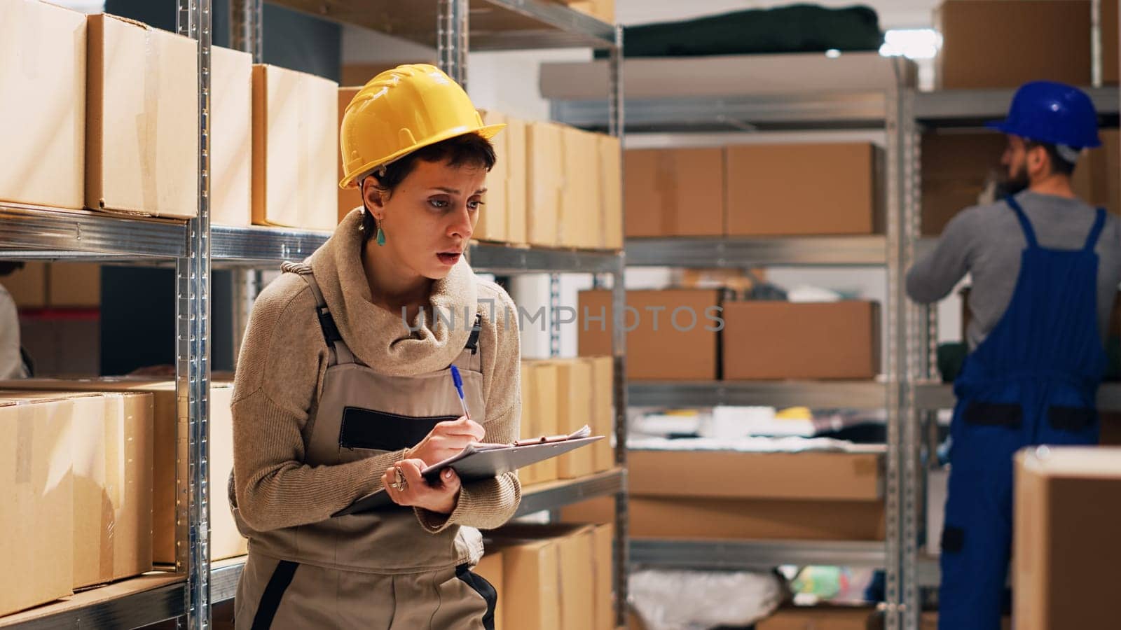 Female supervisor checking number of merchandise in storage room, counting cardboard boxes with products on depot shelves. Woman using clipboard files to look at logistics list. Handheld shot.