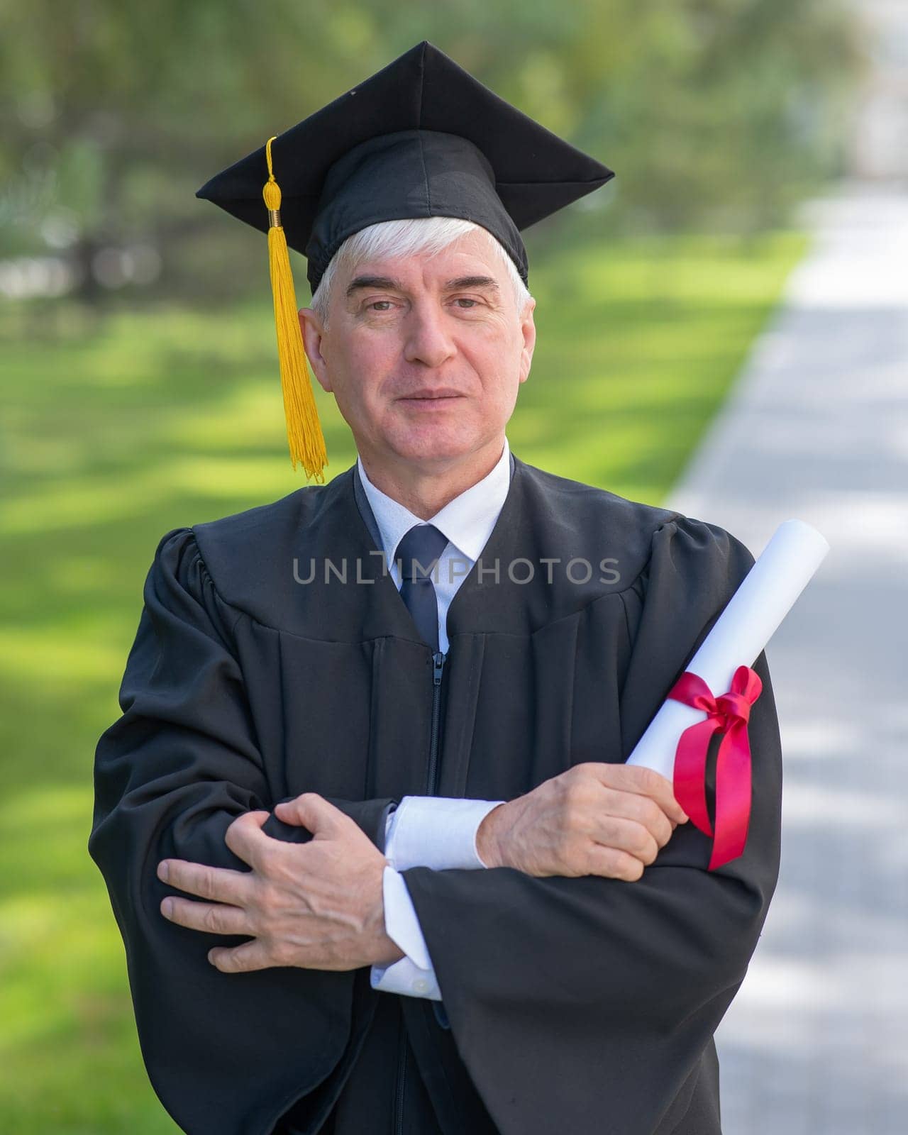 Portrait of an elderly man in a graduation gown and with a diploma in his hands outdoors. Vertical