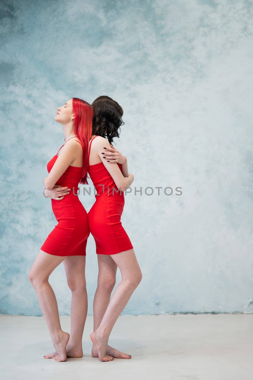 A full-length portrait of two women dressed in identical red dresses and standing back to back. Lesbian intimacy