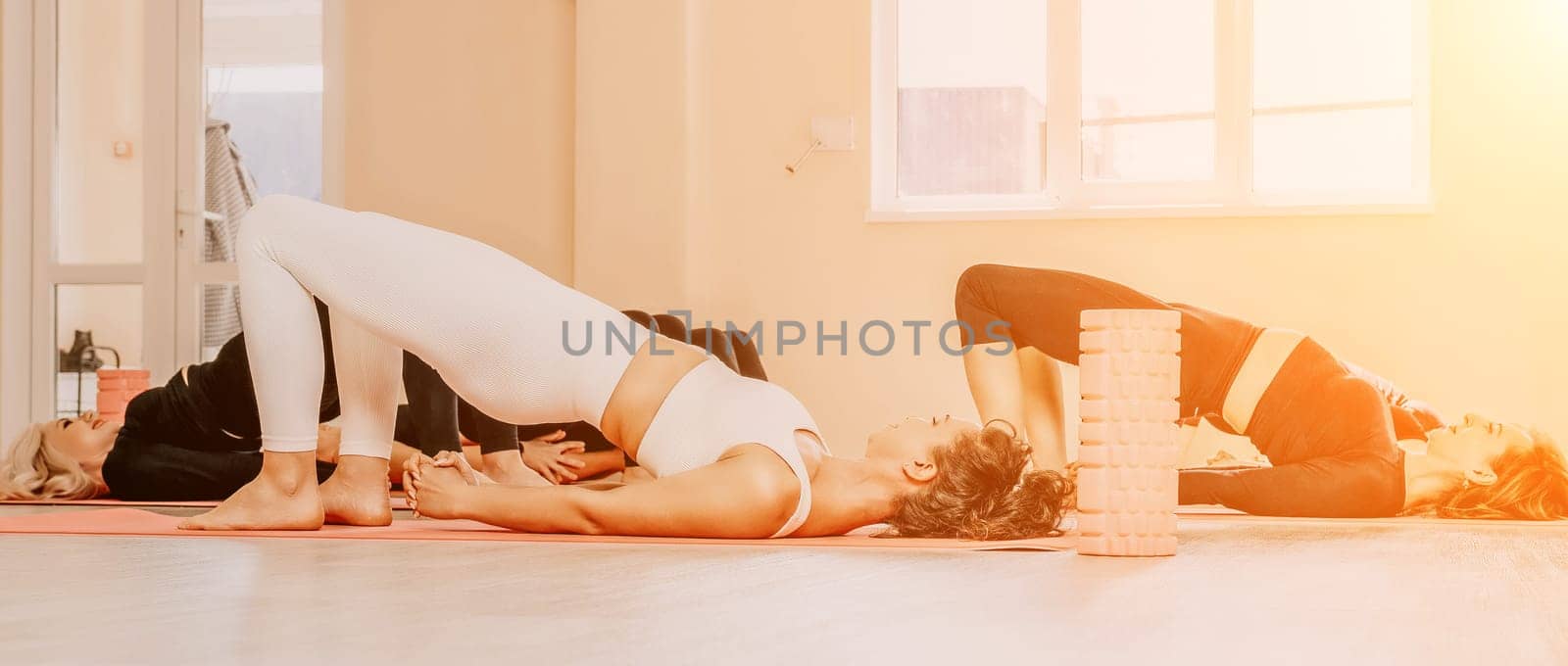 Group of young womans fitness instructor in Sportswear Leggings and Tops, stretching in the gym before pilates, on a yoga mat near the large window on a sunny day, female fitness yoga routine concept.