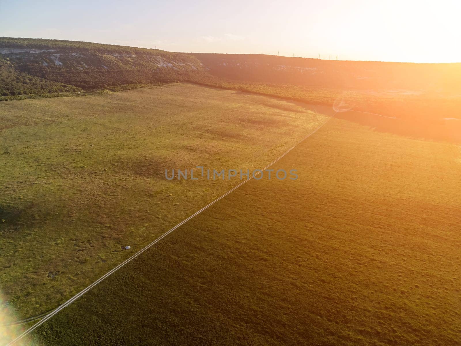 Aerial view on green wheat field, road and hills in countryside. Field of wheat blowing in the wind on sunset. Ears of barley crop in nature. Agronomy, industry and food production. by panophotograph