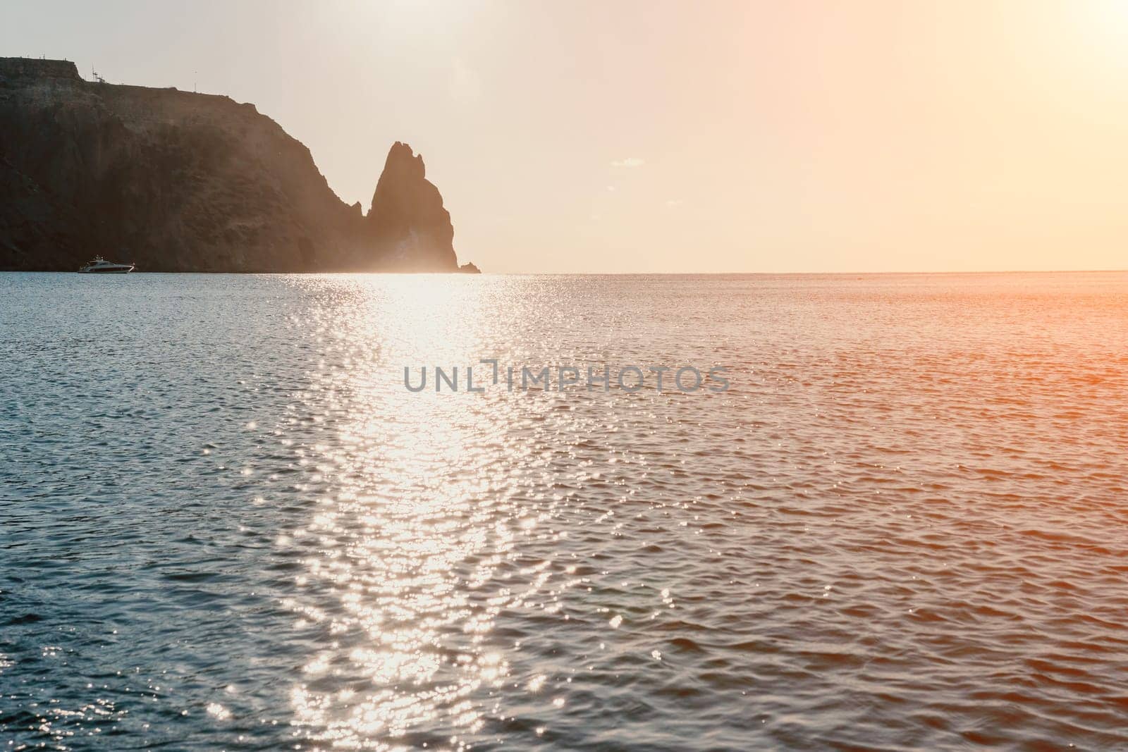 A red burning sunset with the silhouette of a cliff over the sea. Abstract nature summer or spring ocean sea background. Small waves on golden warm water surface with bokeh lights from sun.