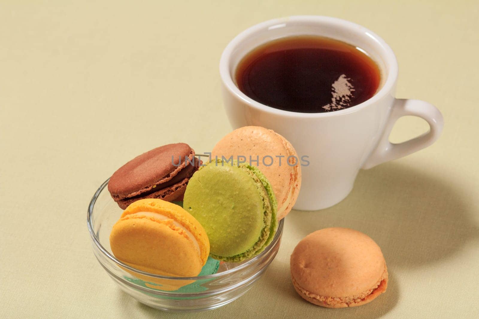 Tasty macaroons cakes of different color in glass bowl and cup of coffee on beige background.