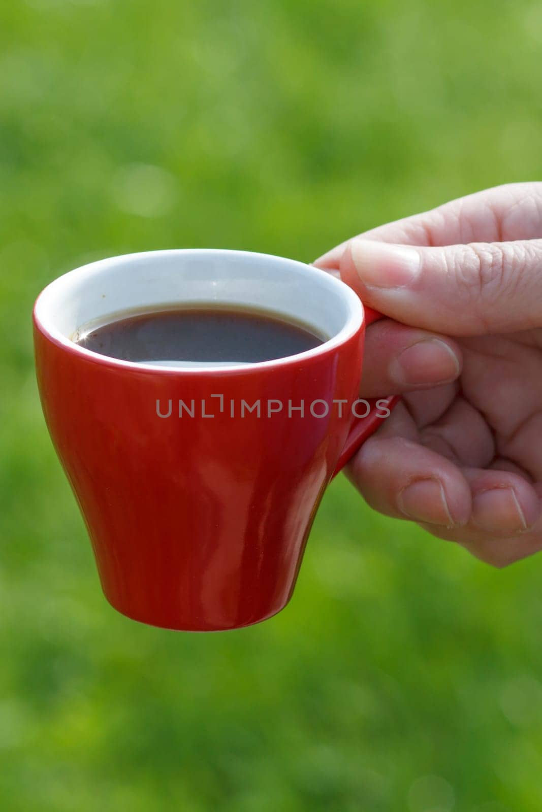 Female hand holds a red porcelain cup of coffee with green grass on the background. Selective focus on cup.
