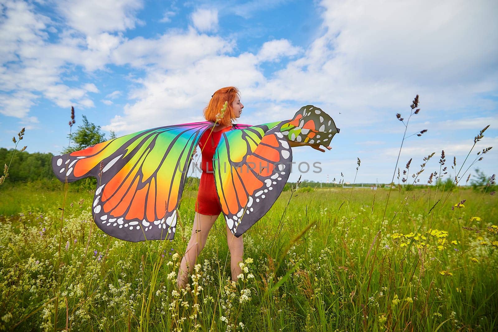 Adult girl with red hair and butterfly wings having fun and joy in meadow or field with grass, flowers on sunny summer day