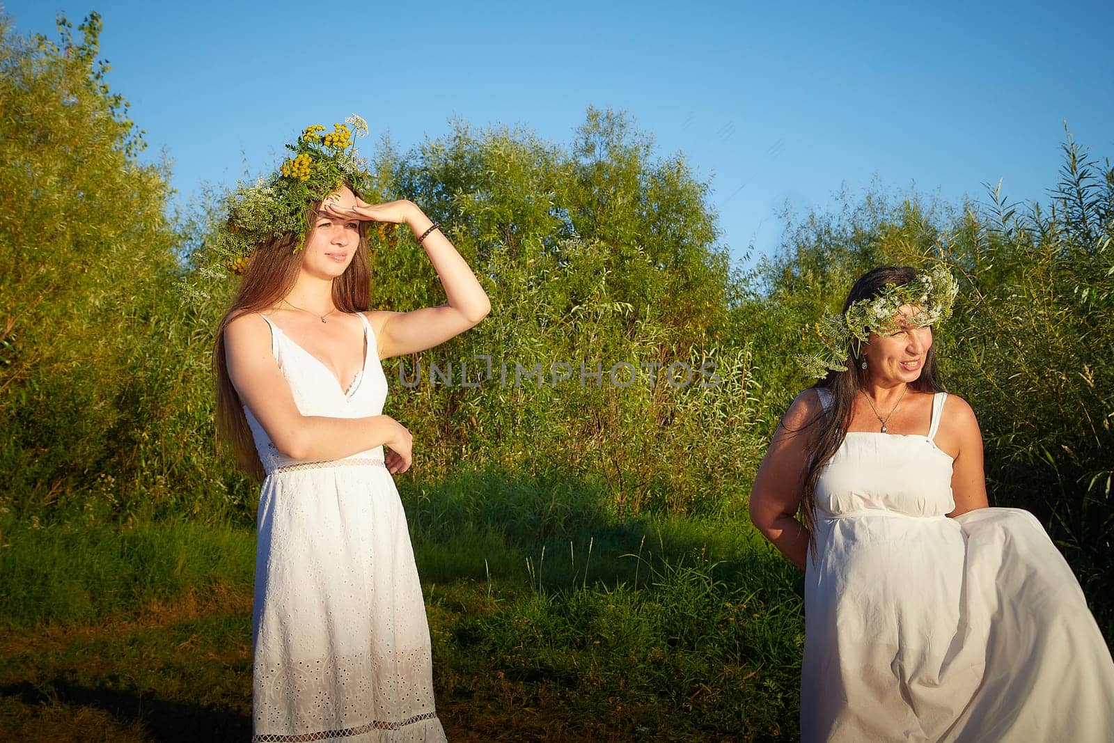 Adult mature and young brunette women in white sundresses and wreaths of flowers in summer by water of river or lake at sunset. Girl mother and daughter Celebrating pagan holiday of Ivan Kupala