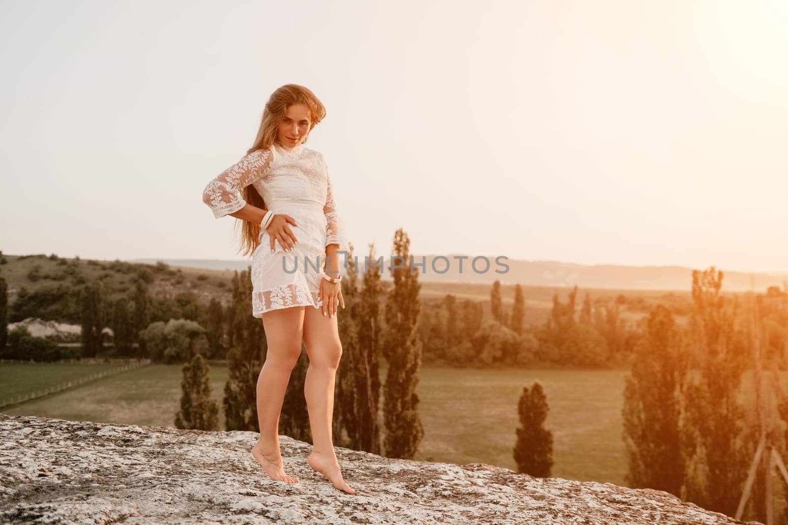 Romantic beautiful bride in white dress posing with sea and mountains in background. Stylish bride standing back on beautiful landscape of sea and mountains on sunset