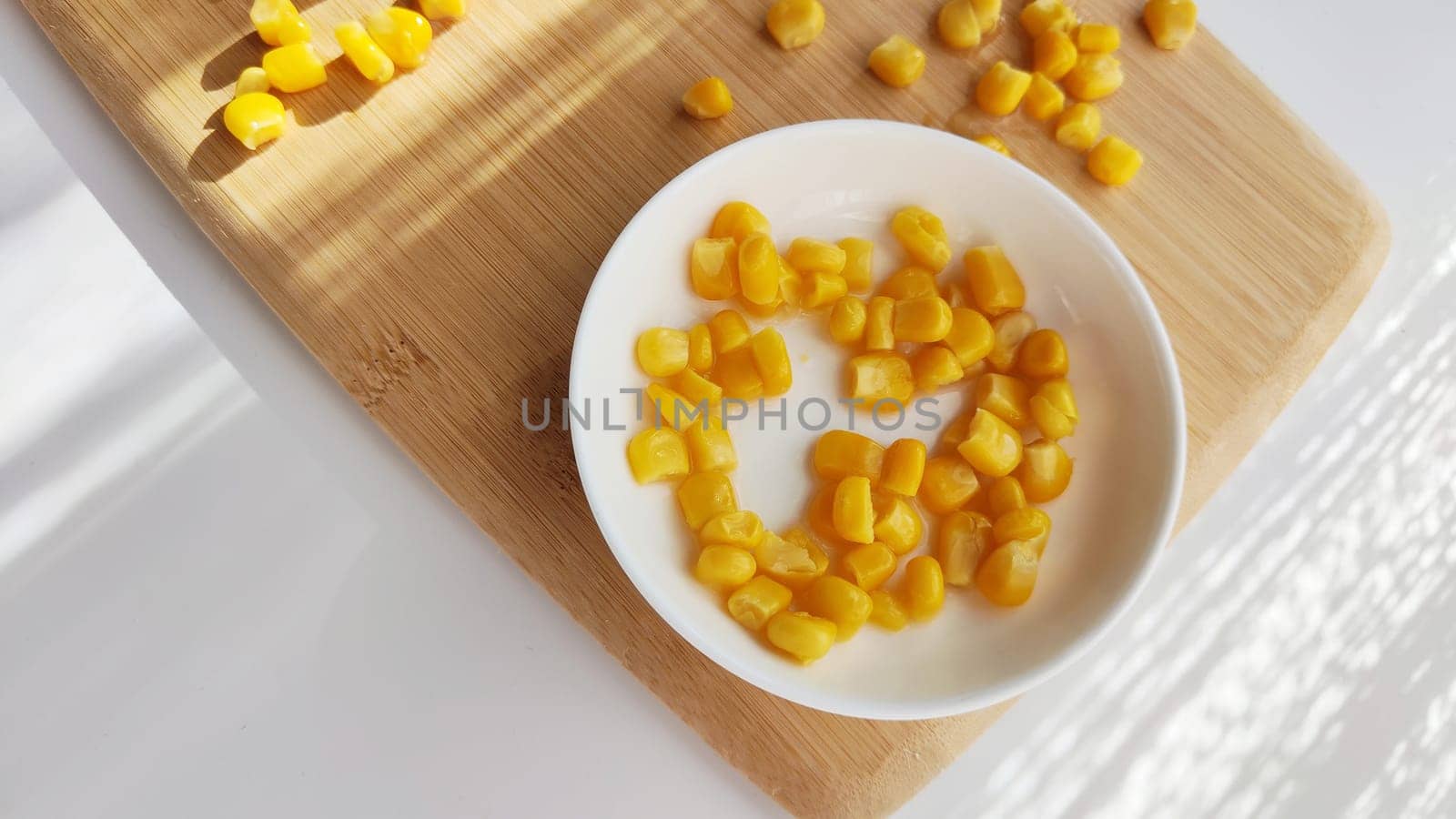 Lot of pieces of canned yellow corn on plate which is on wooden bamboo cutting board on white background. Concept of cooking and delicious healthy food