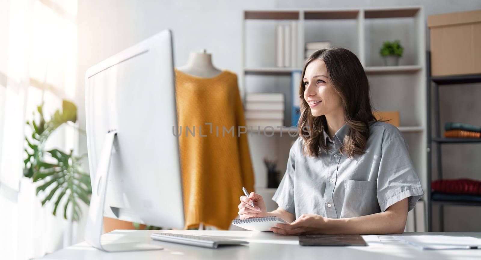 Young attractive female fashion designer sketching idea on paper at desk, working with a laptop at home.