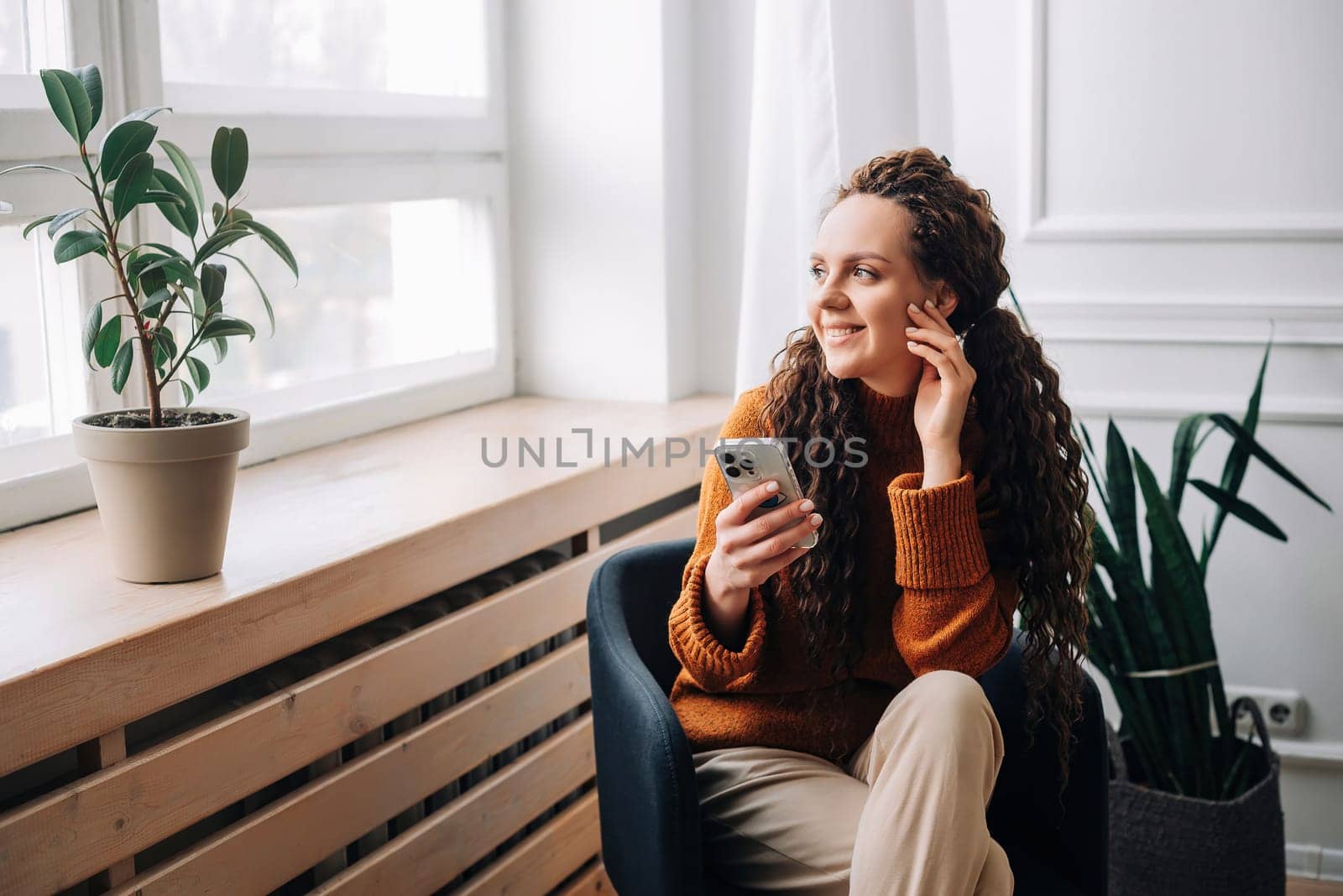 Young woman enjoying smartphone apps and texting while sitting on a chair. Mobile phone user: woman checking apps, messaging, and shopping from home. Relaxed woman browsing modern apps.
