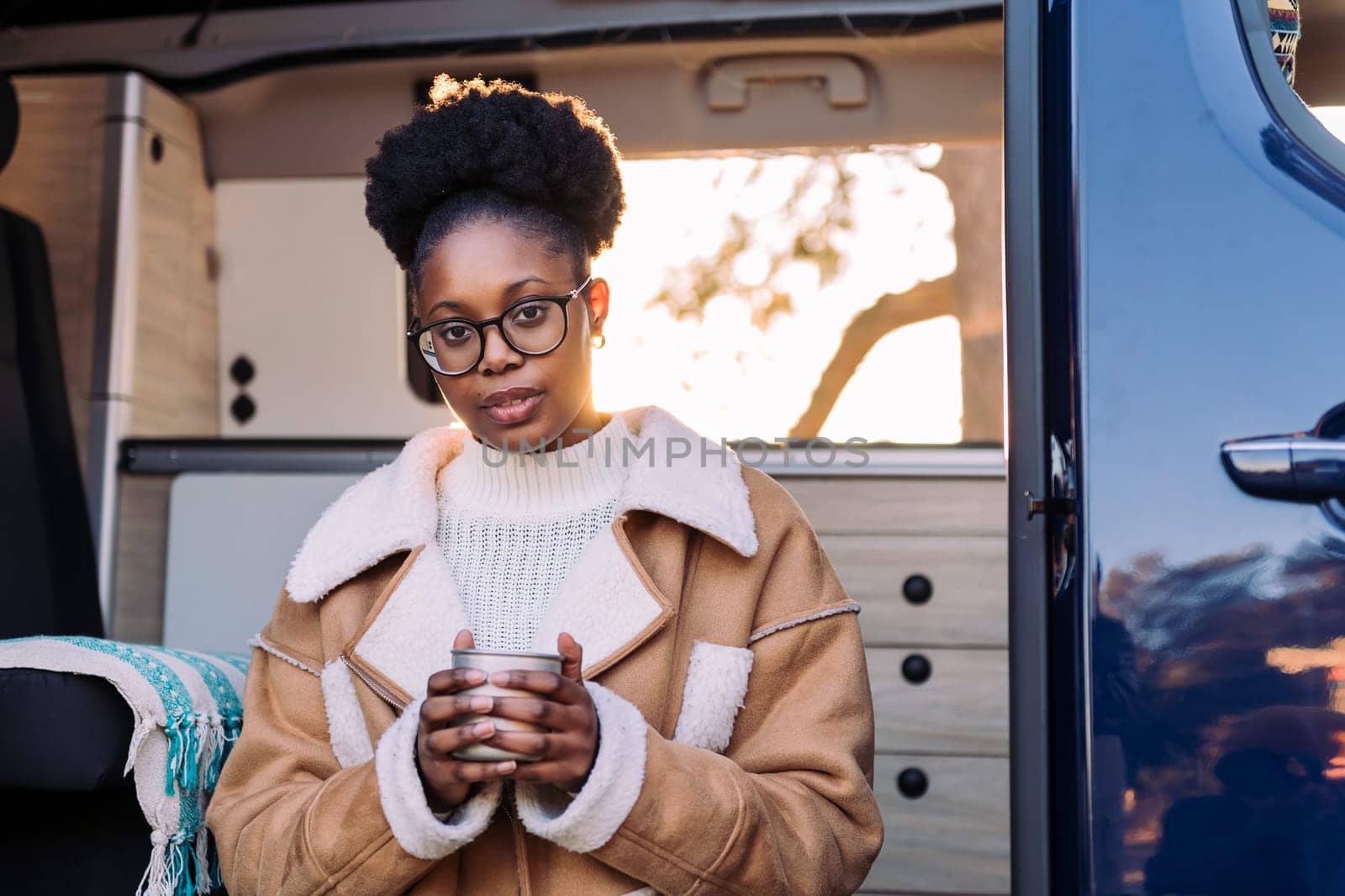 young african woman relaxing and enjoying a cup of a hot drink at sunset in a camper van, concept of van life and weekend getaway, copy space for text