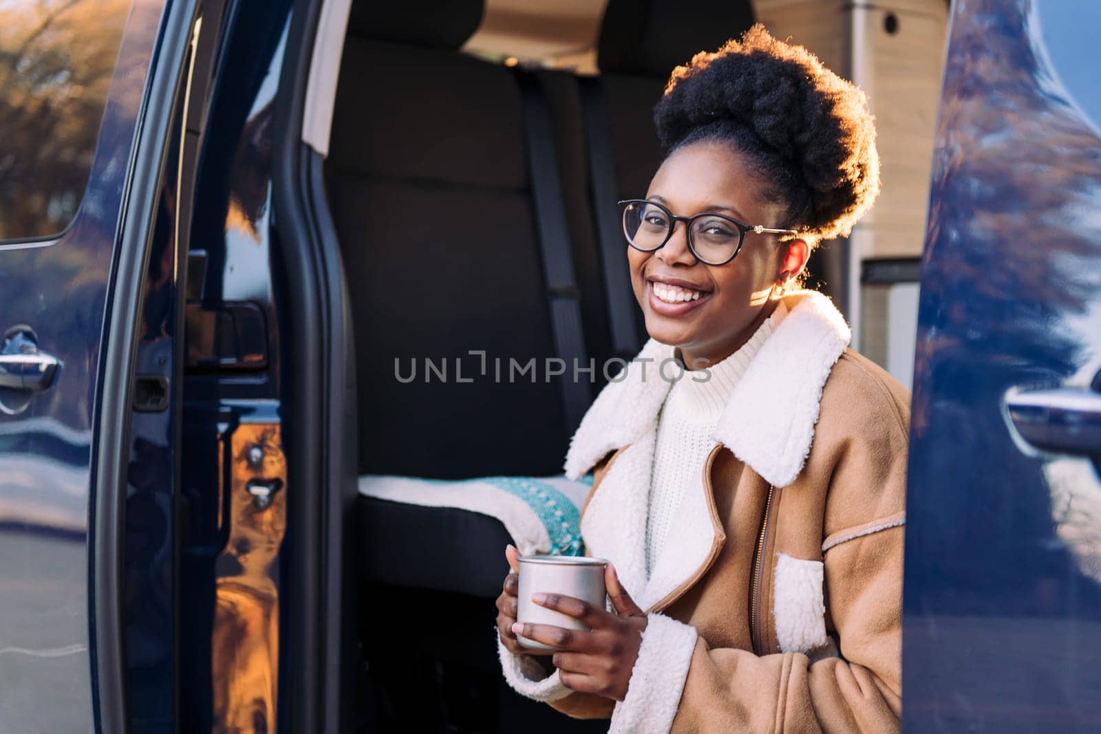 portrait of a young african woman smiling happy in a camper van looking at camera with a mug in her hand, concept of van life and weekend getaway, copy space for text