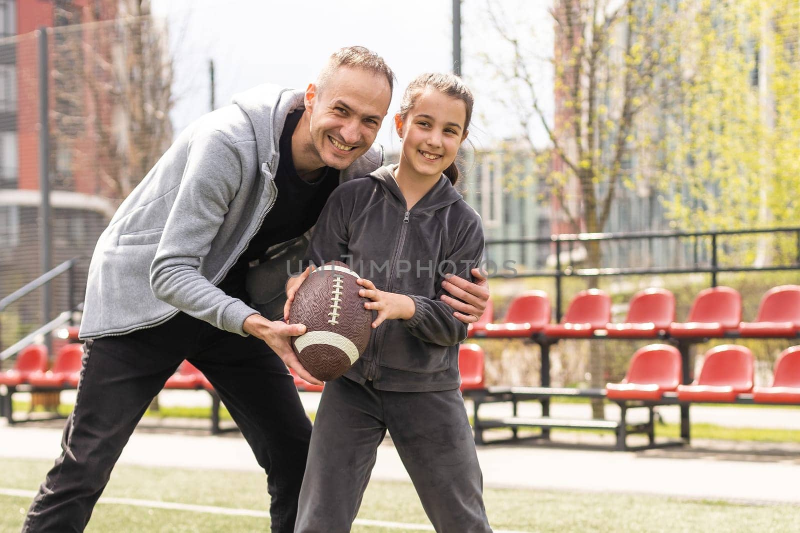 Handsome dad with his little cute daughter are having fun and playing American football on green grassy lawn.