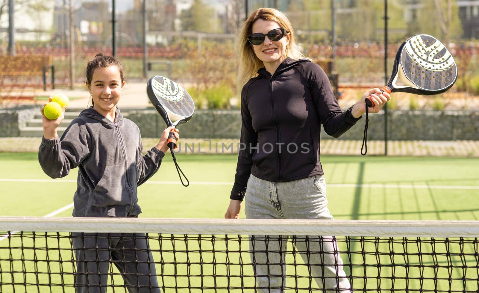 Full body of positive mother and daughter standing on court with padel rackets during break and looking at camera