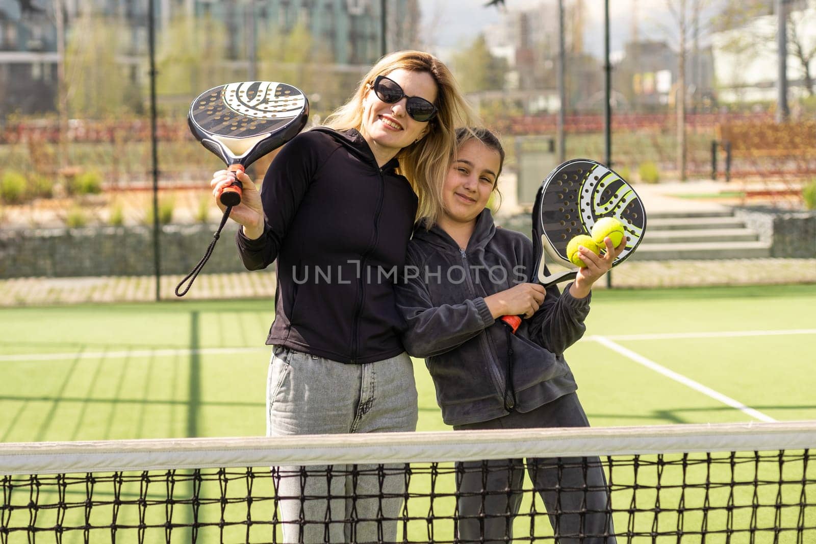 Full body of positive mother and daughter standing on court with padel rackets during break and looking at camera