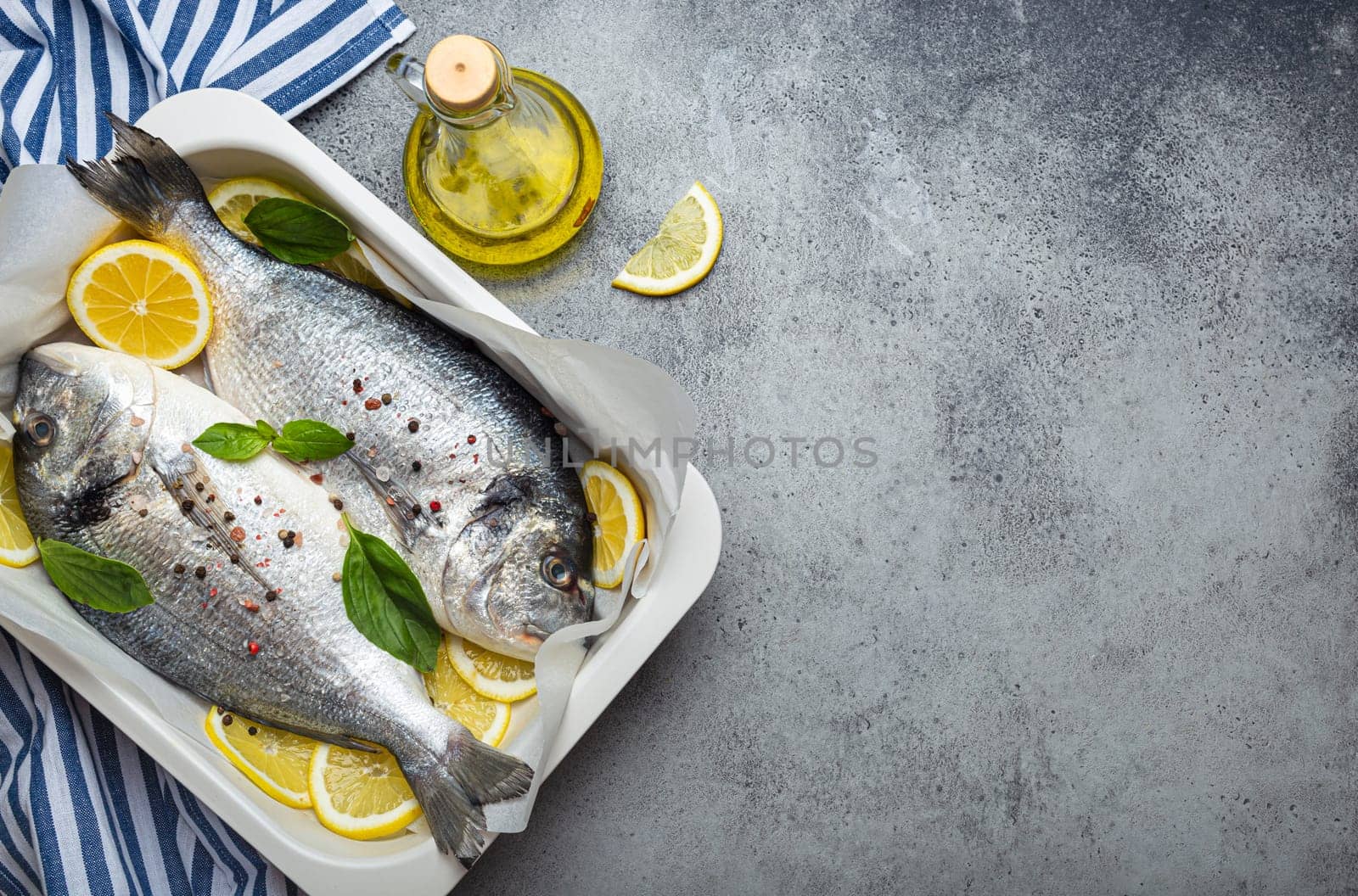 Raw fish dorado in casserole dish with ingredients lemon, fresh basil, bottle of olive oil on wooden cutting board on rustic stone background top view, cooking healthy fish dorado. Copy space