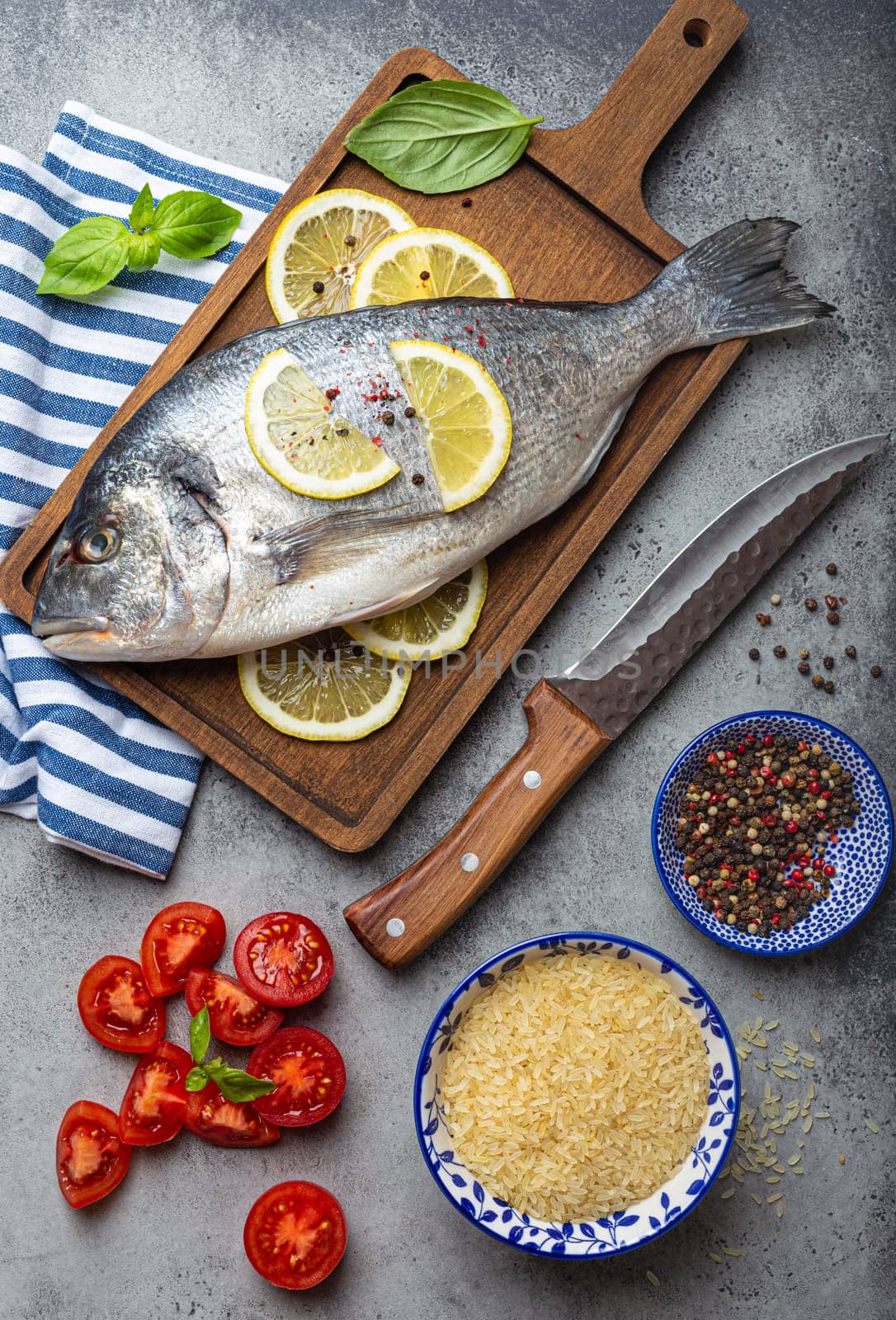 Raw fish dorado with ingredients lemon, fresh basil, cut cherry tomatoes, uncooked rice on wooden cutting board with knife on rustic stone background top view, cooking healthy fish dorado by its_al_dente