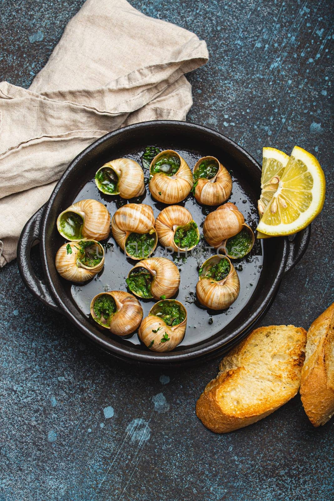 Escargots de Bourgogne Snails with Garlic Butter and Parsley in black cast iron pan with Lemon and Toasted Baguette Slices on rustic stone background top view, traditional French Delicacy . by its_al_dente