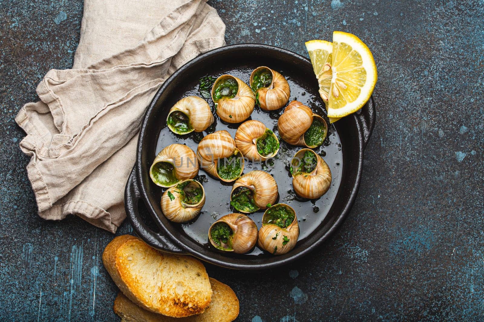 Escargots de Bourgogne Snails with Garlic Butter and Parsley in black cast iron pan with Lemon and Toasted Baguette Slices on rustic stone background top view, traditional French Delicacy .