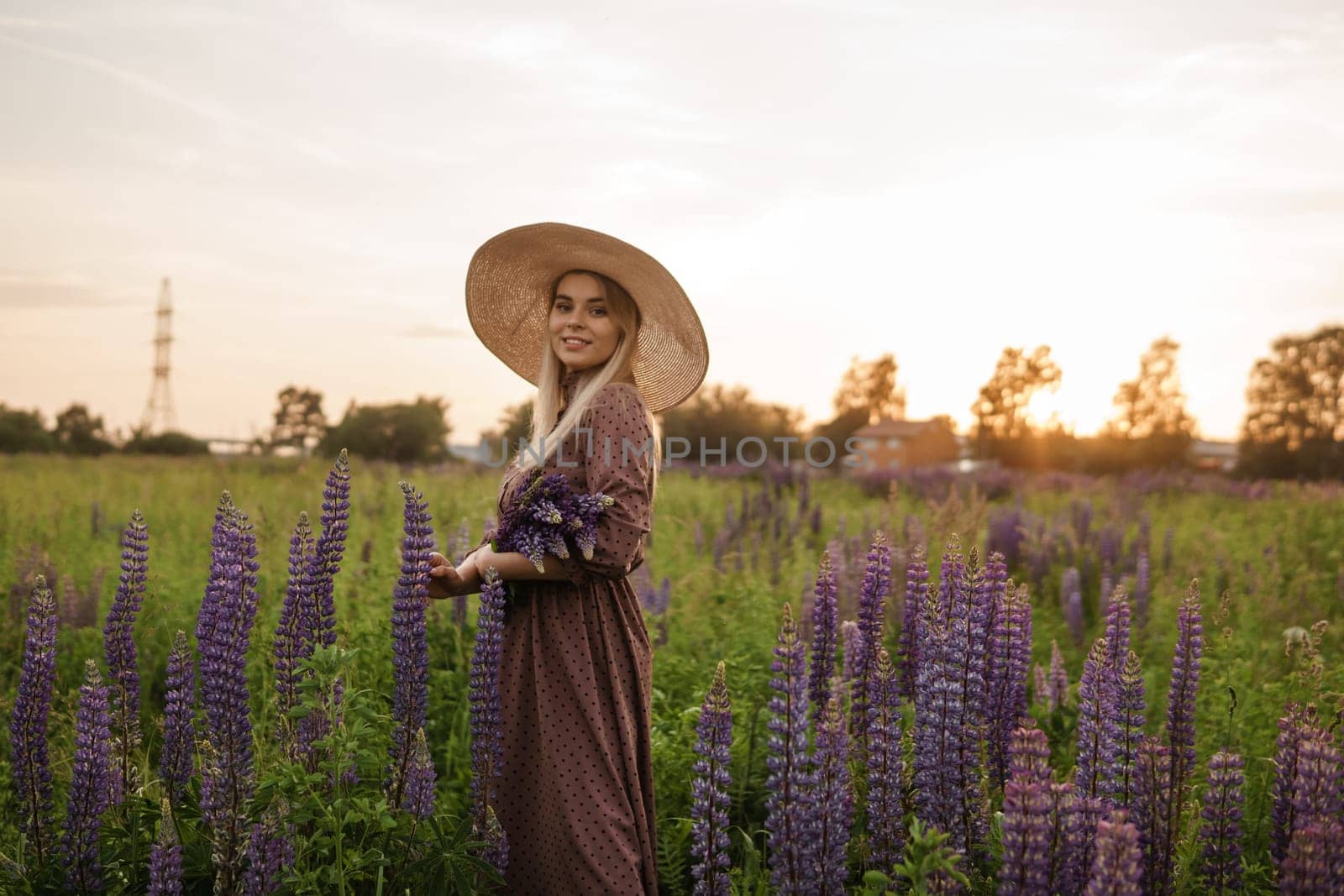 A beautiful woman in a straw hat walks in a field with purple flowers. A walk in nature in the lupin field.