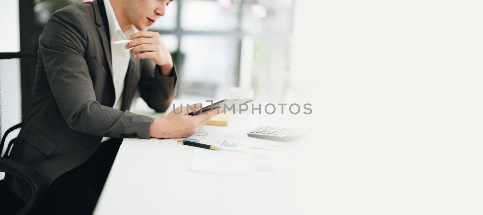 Portrait of a young Asian man showing a smiling face as she uses his phone, computer and financial documents on her desk in the early morning hours.
