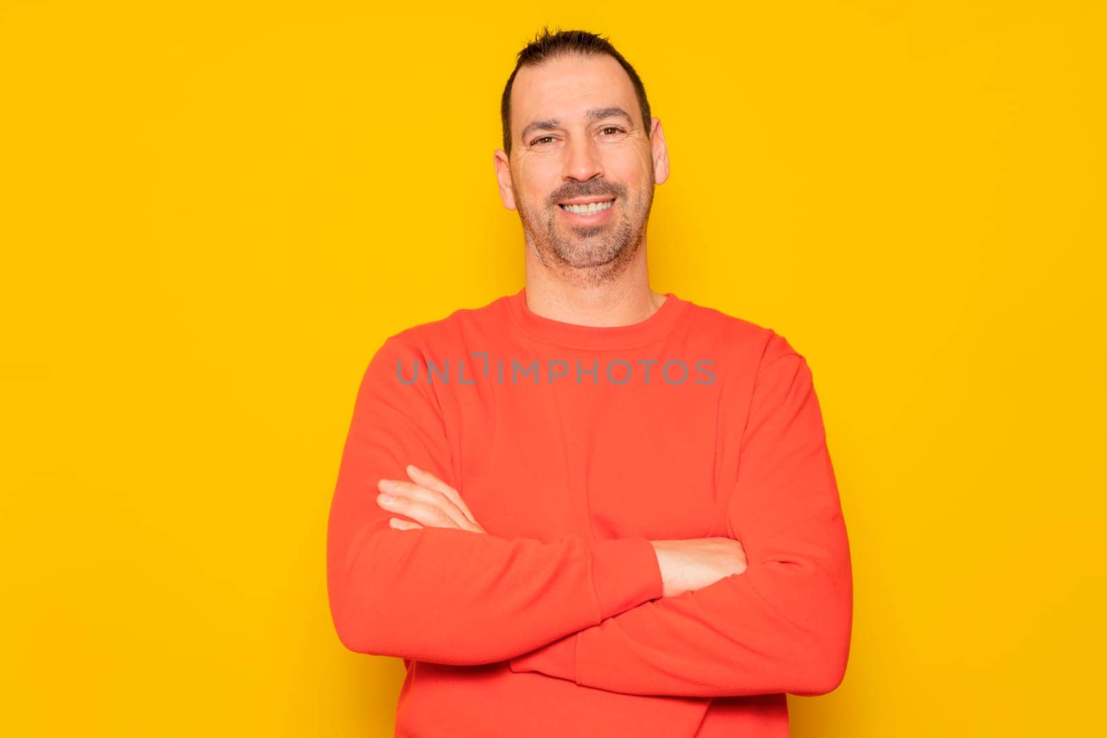 Portrait of a smart, cheerful and attractive bearded Hispanic man in his 40s, arms crossed, isolated over yellow colored background