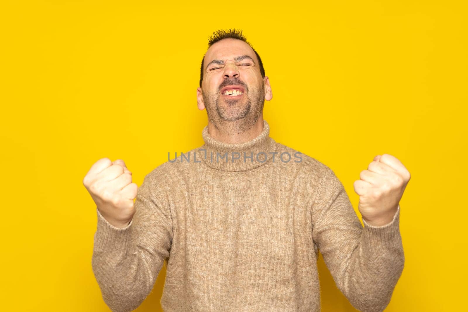 Bearded Hispanic man in his 40s wearing a beige turtleneck energetically celebrating a big win with raised fists, isolated on yellow studio background