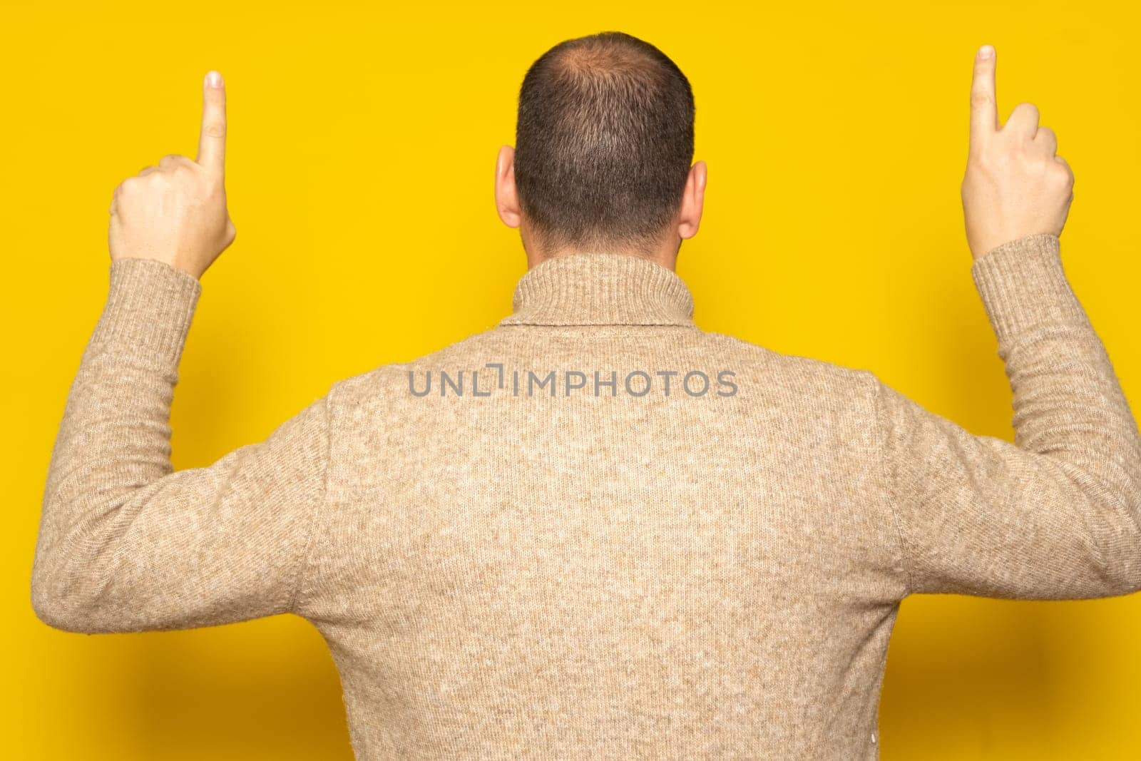 Hispanic man in casual clothing posing backwards pointing up with his index fingers, isolated on yellow studio background