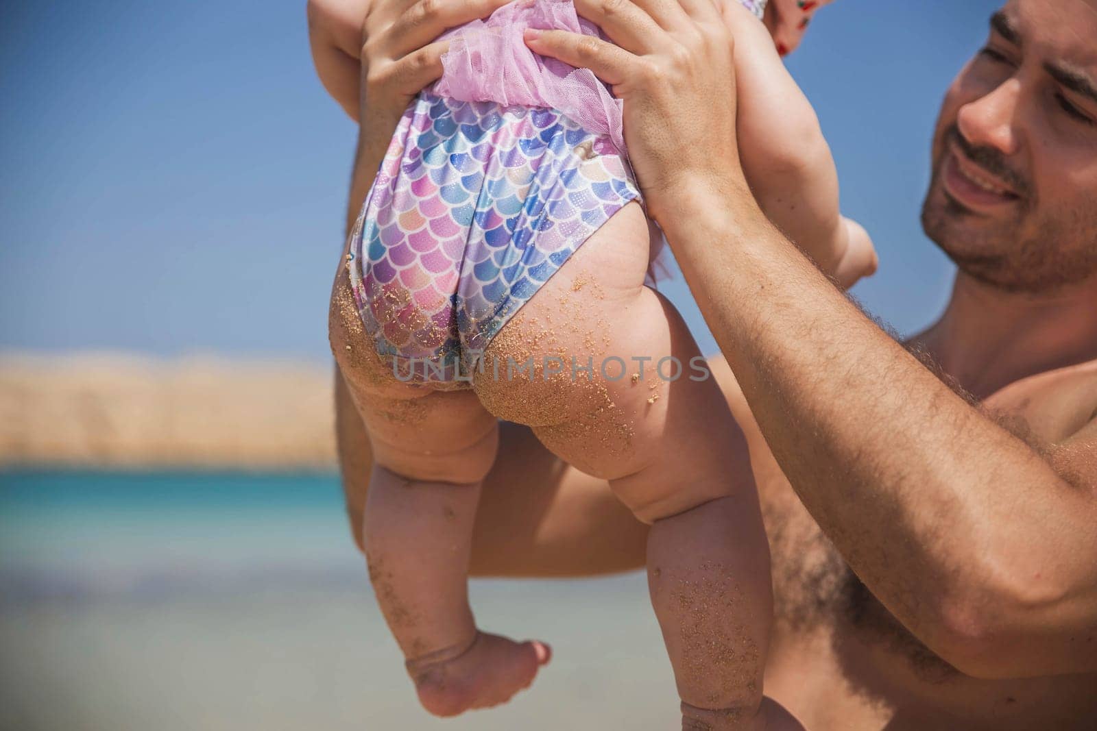 father holds newborn baby in a fish swimsuit on the beach.