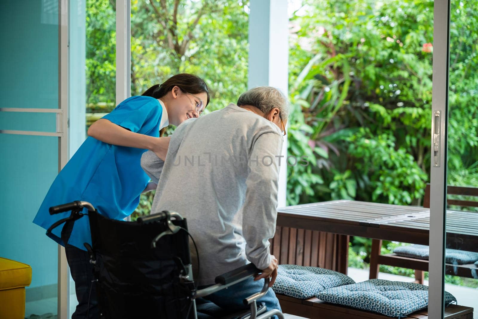 Young Asian nurse is supporting elderly man sitting in wheelchair walking to looking out window on garden and breathe fresh air, pension retirement center, senior man at home, healthcare concept