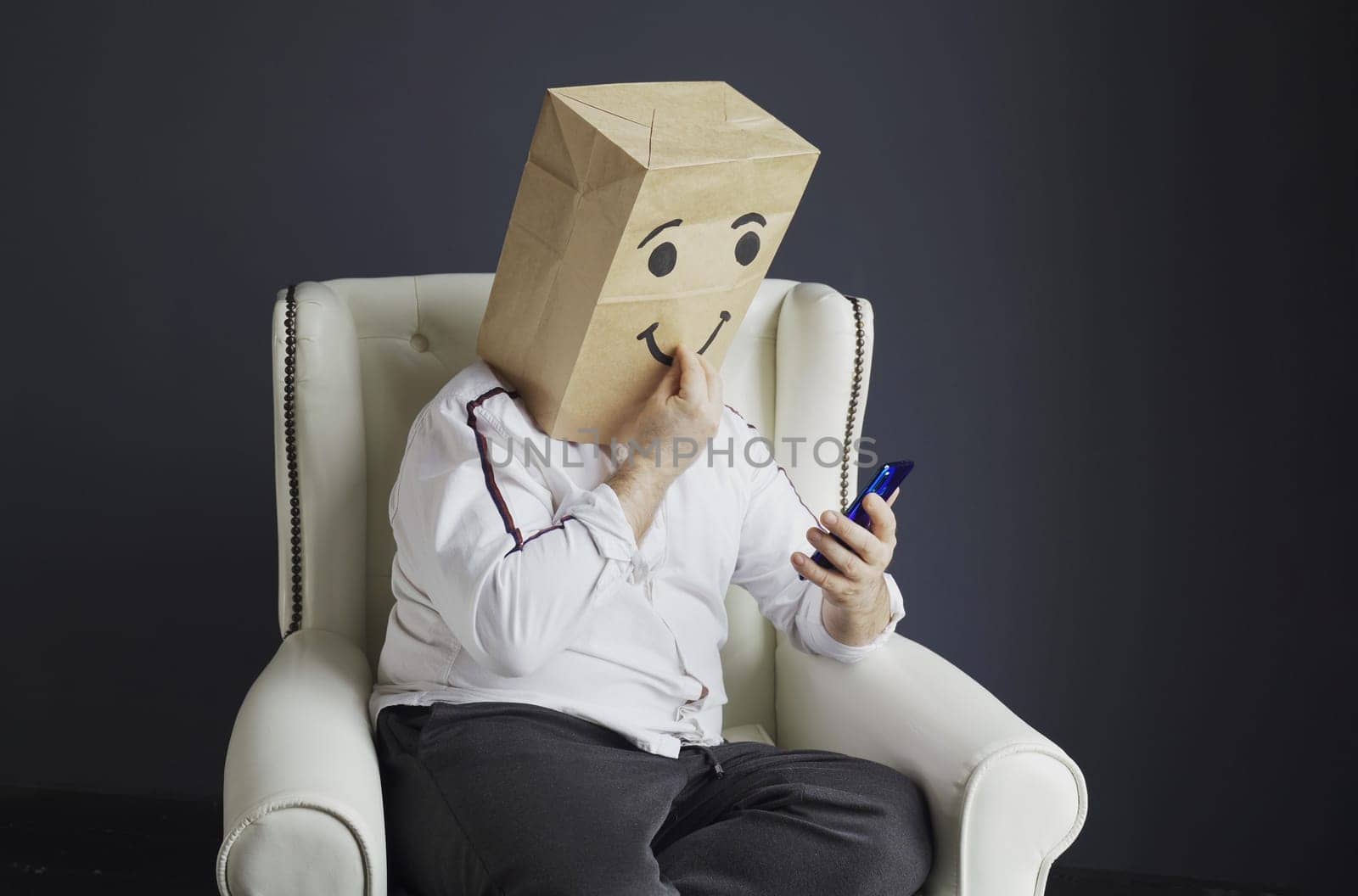 A man in a white shirt with a paper bag on his head, with a smiley face drawn, sits in a chair and talks on a video call on a smartphone, sends air kisses.