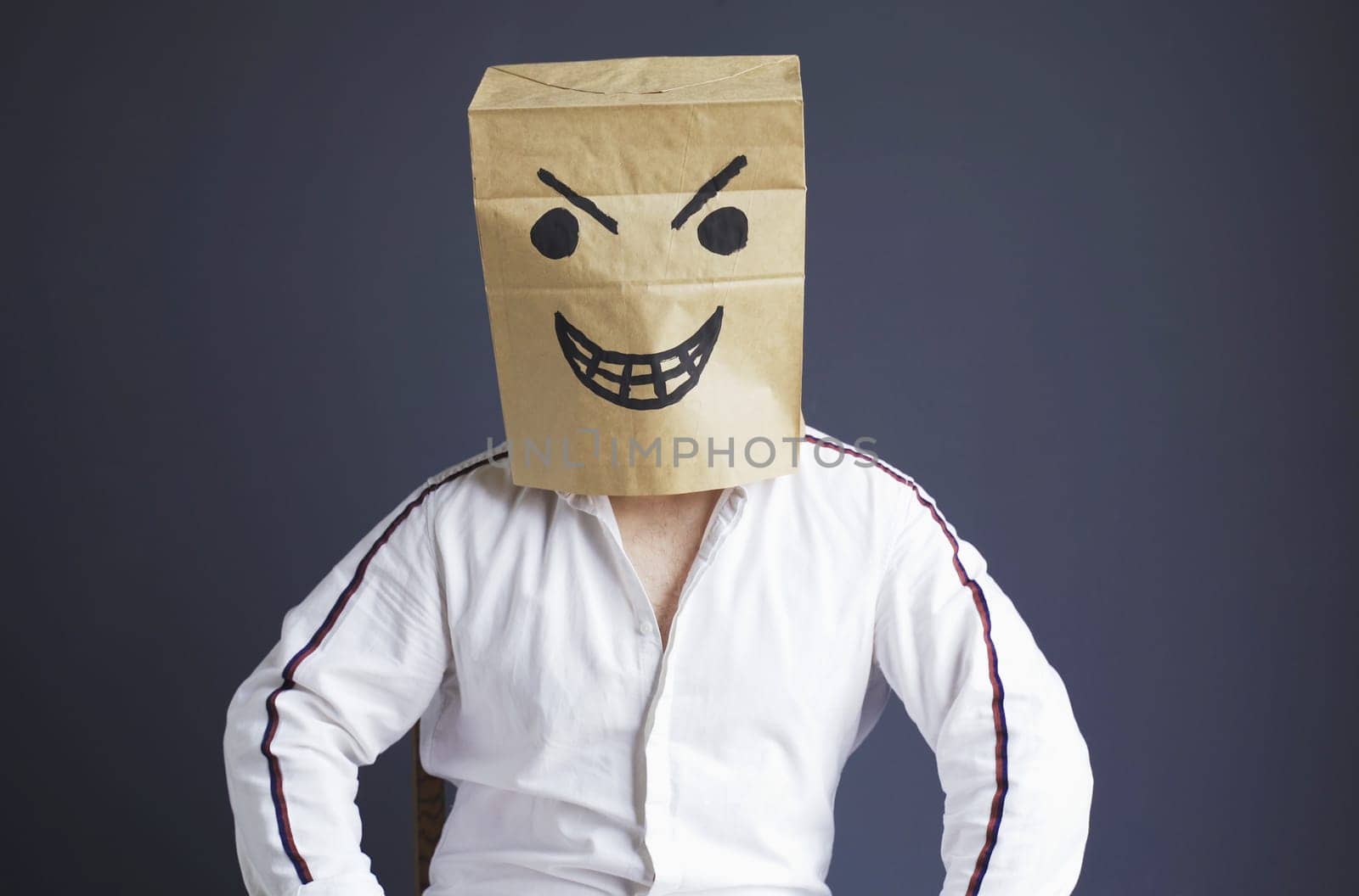 A man sits on a chair with a paper bag on his head, with a drawn angry smiley. Emotions and gestures.