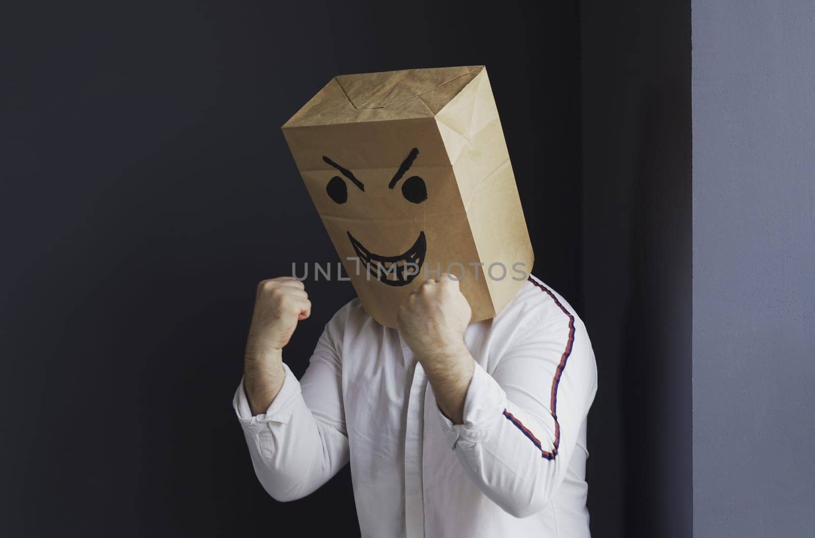 A man in a white shirt with a paper bag with an angry emoticon on his head gesticulates aggressively with his hands. Emotions and anger