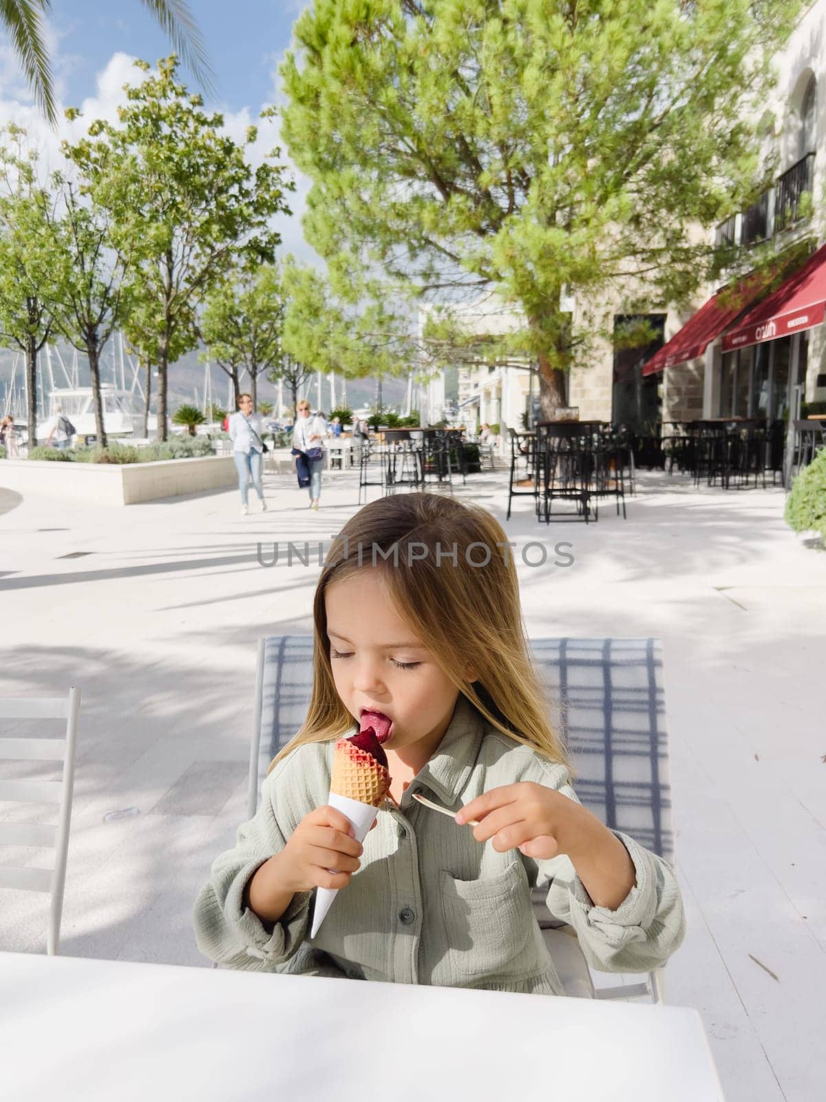 Little girl licks popsicles in a waffle cone while sitting on a chair in a street cafe. High quality photo