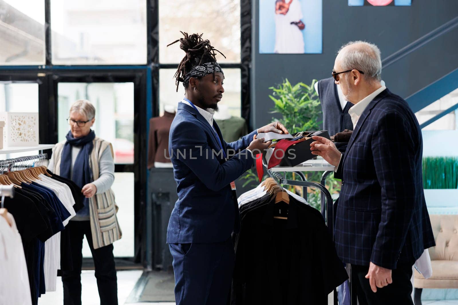 African american employee helping customer with right tie, analyzing fabric in modern boutique. Elderly client wearing stylish suit shopping for casual wear and fashionable merchandise in showroom