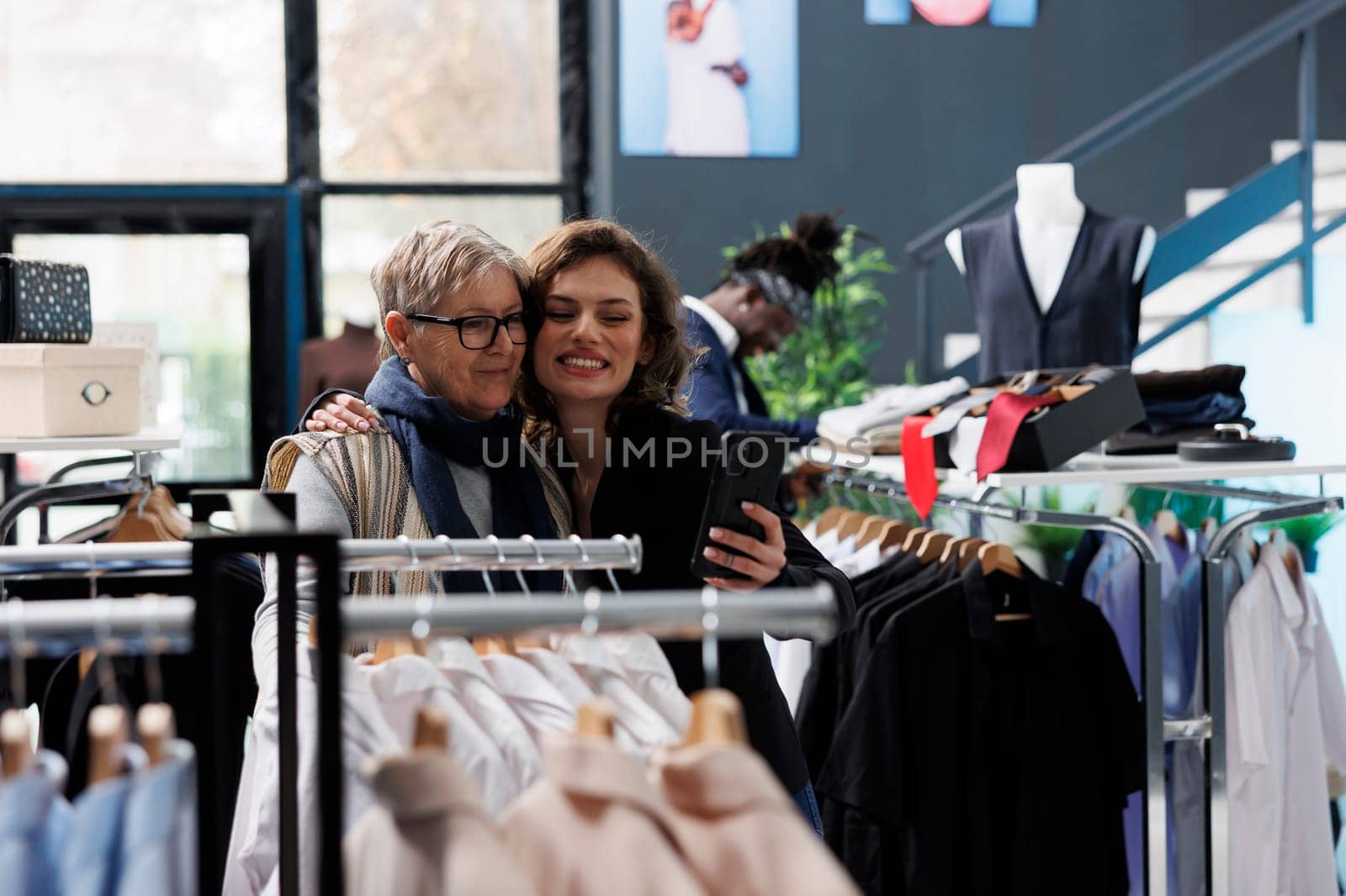 Cheerful women smiling at camera taking selfie with white shirt using phone, having fun in shopping centre. Showroom clients buying fashionable clothes and merchandise in modern boutique