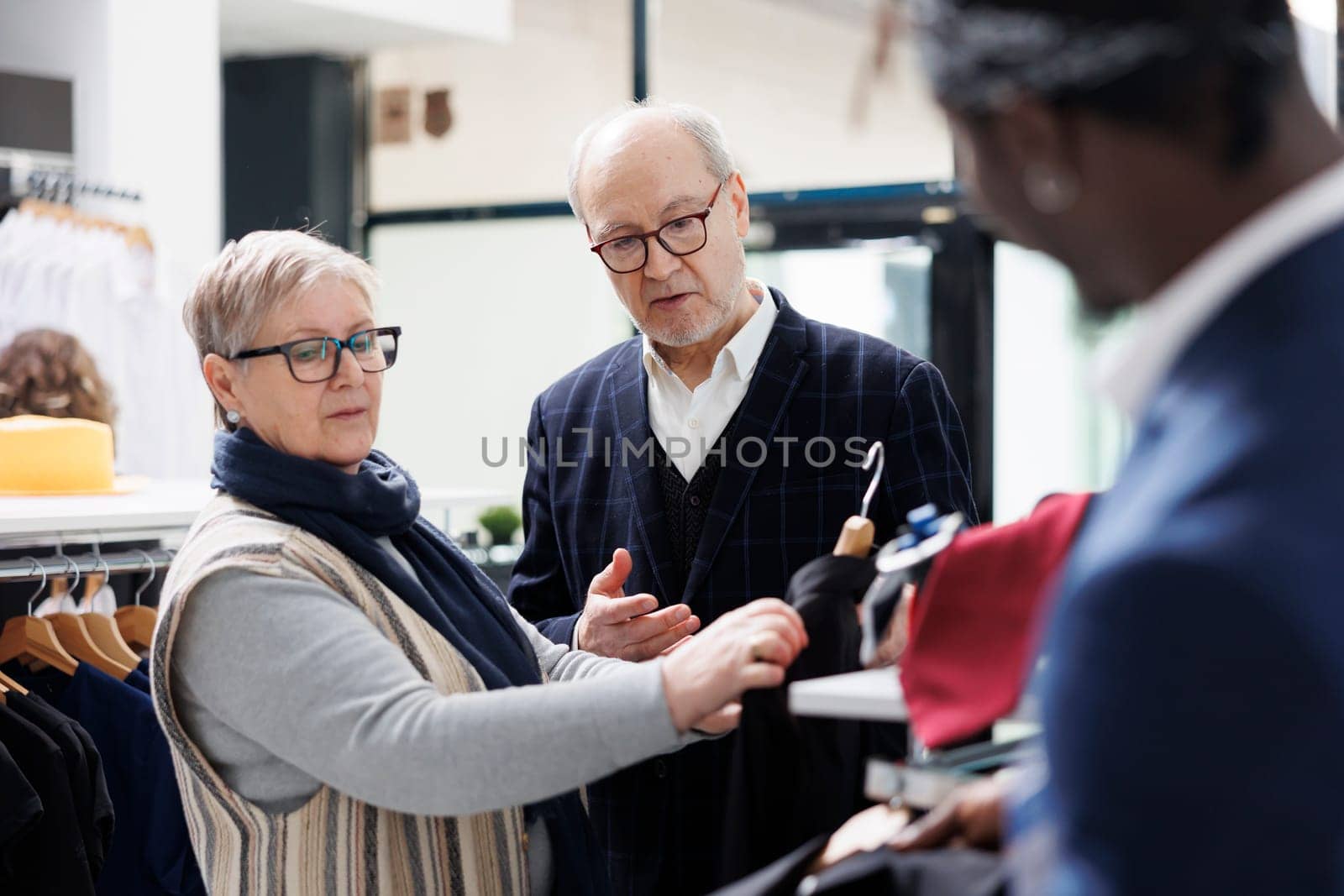 Senior couple looking to buy casual wear, asking store worker about clothes material in modern boutique. Elderly man buying fashionable merchandise and stylish accessories in shopping centre