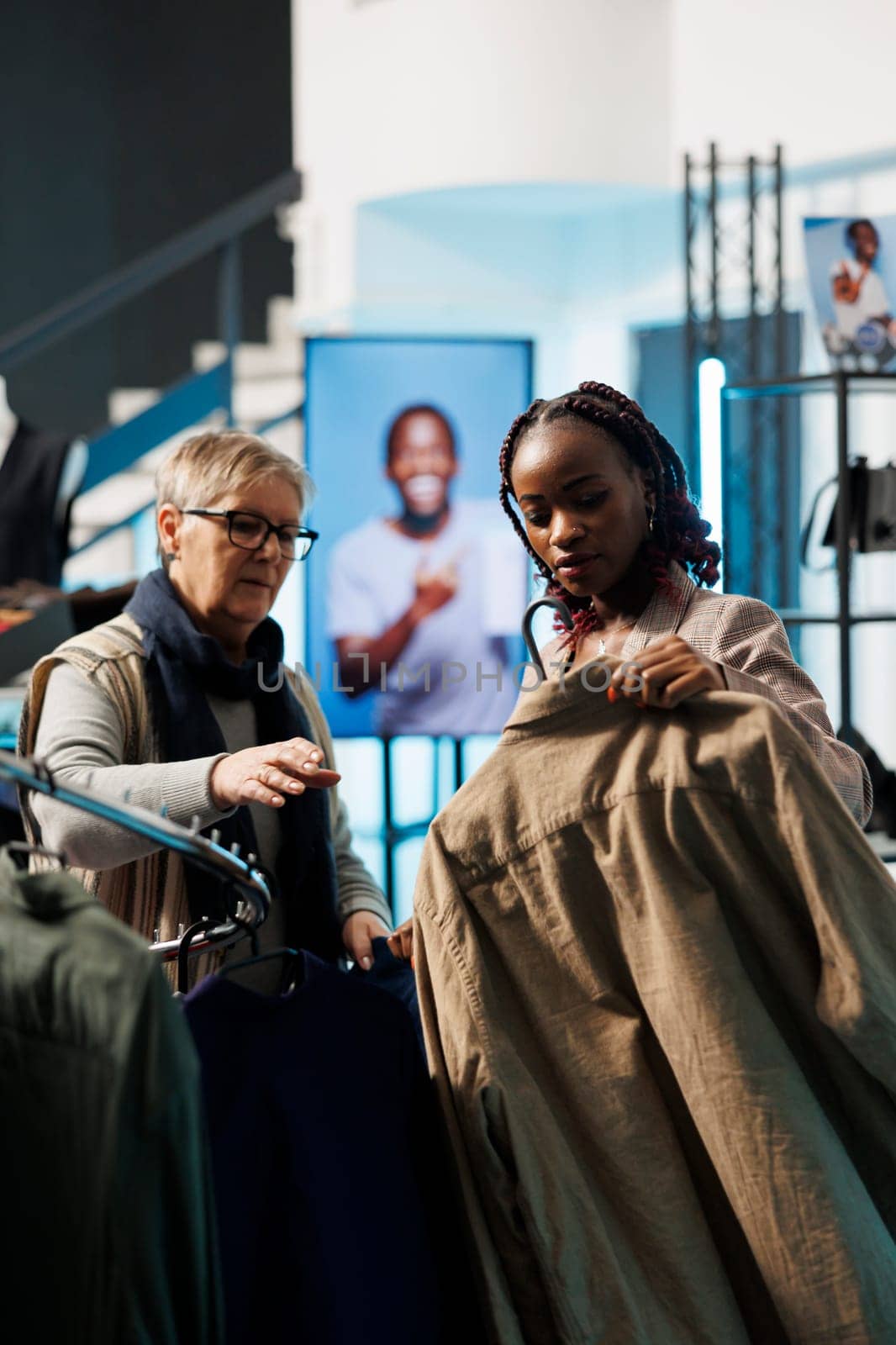 African american employee helping elderly client choosing brown shirt for husband in modern boutique. Senior woman shopping for casual wear, buying fashionable merchandise in clothing store