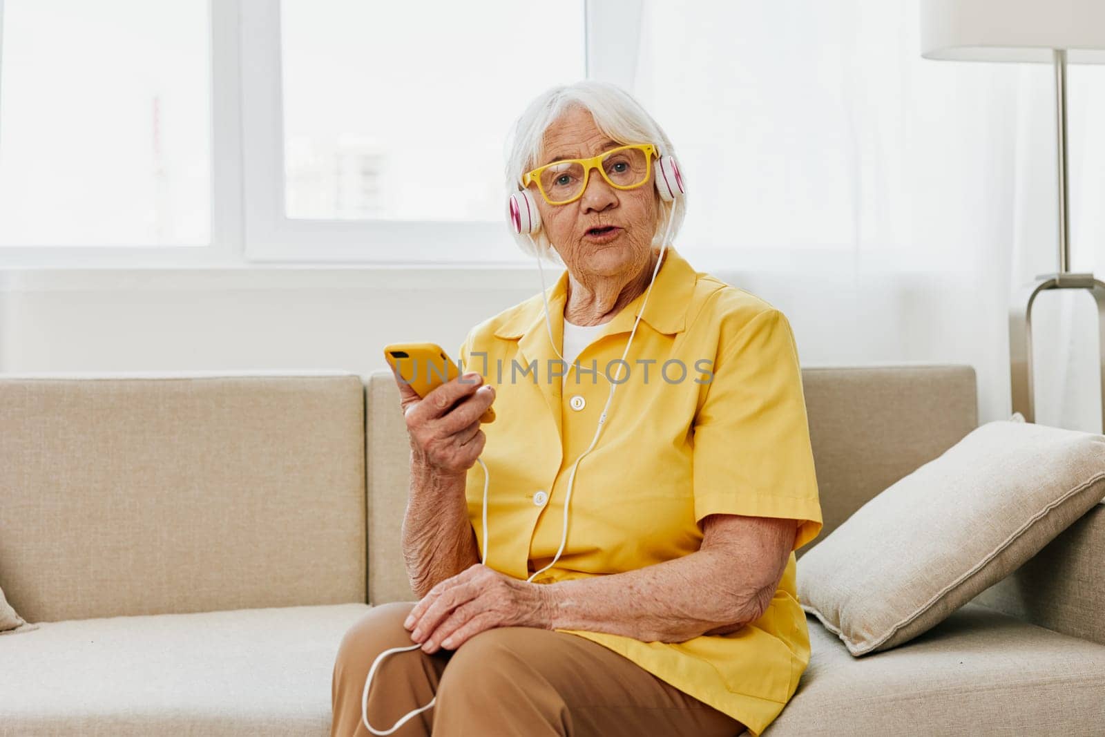 Happy elderly woman looking into her phone video call with headphones smile, technology for communication, bright modern interior, lifestyle online communication by SHOTPRIME