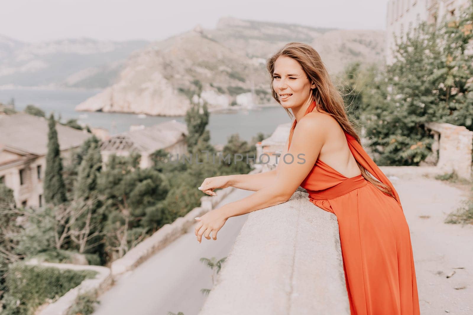 Woman travel portrait. Happy woman with long hair looking at camera and smiling. Close up portrait cute woman in a res long dress posing on backdrop of old travel city near sea by panophotograph