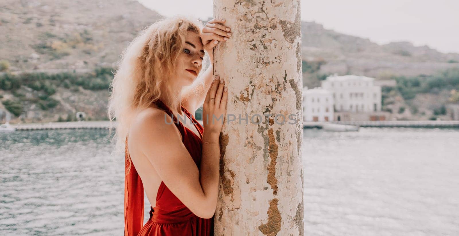 Redhead woman portrait. Curly redhead young caucasian woman with freckles looking at camera and smiling. Cute woman close up portrait in a red long dress posing on sea and travel city background by panophotograph