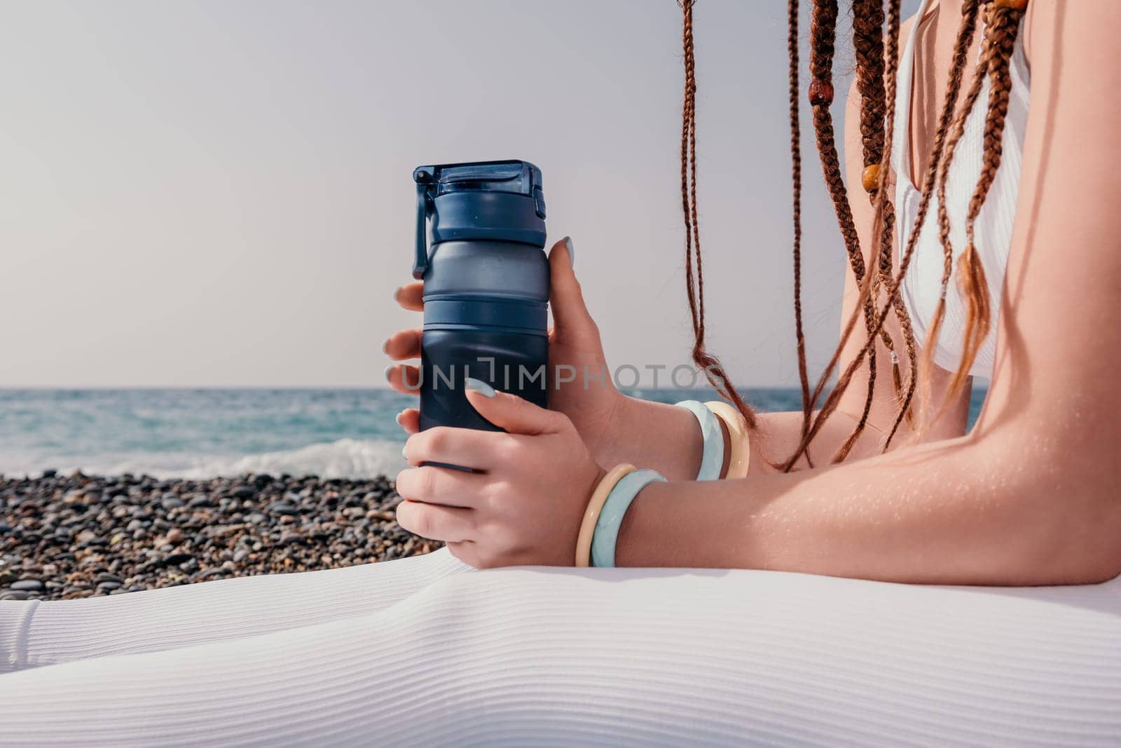 Woman sea yoga. Thermo mug in the hands of woman with braids dreadlocks in white leggings and tops doing stretching pilates on yoga mat near sea. Female fitness yoga routine concept. Healthy lifestyle by panophotograph