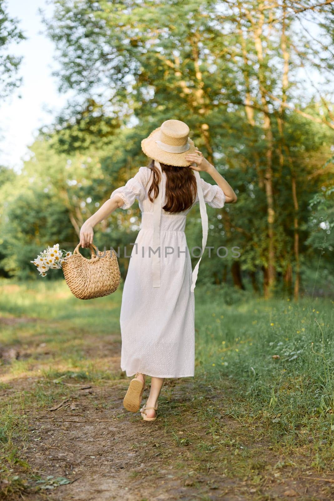 a woman in a long summer dress walks along a forest path holding a wicker hat on her head with her hand by Vichizh