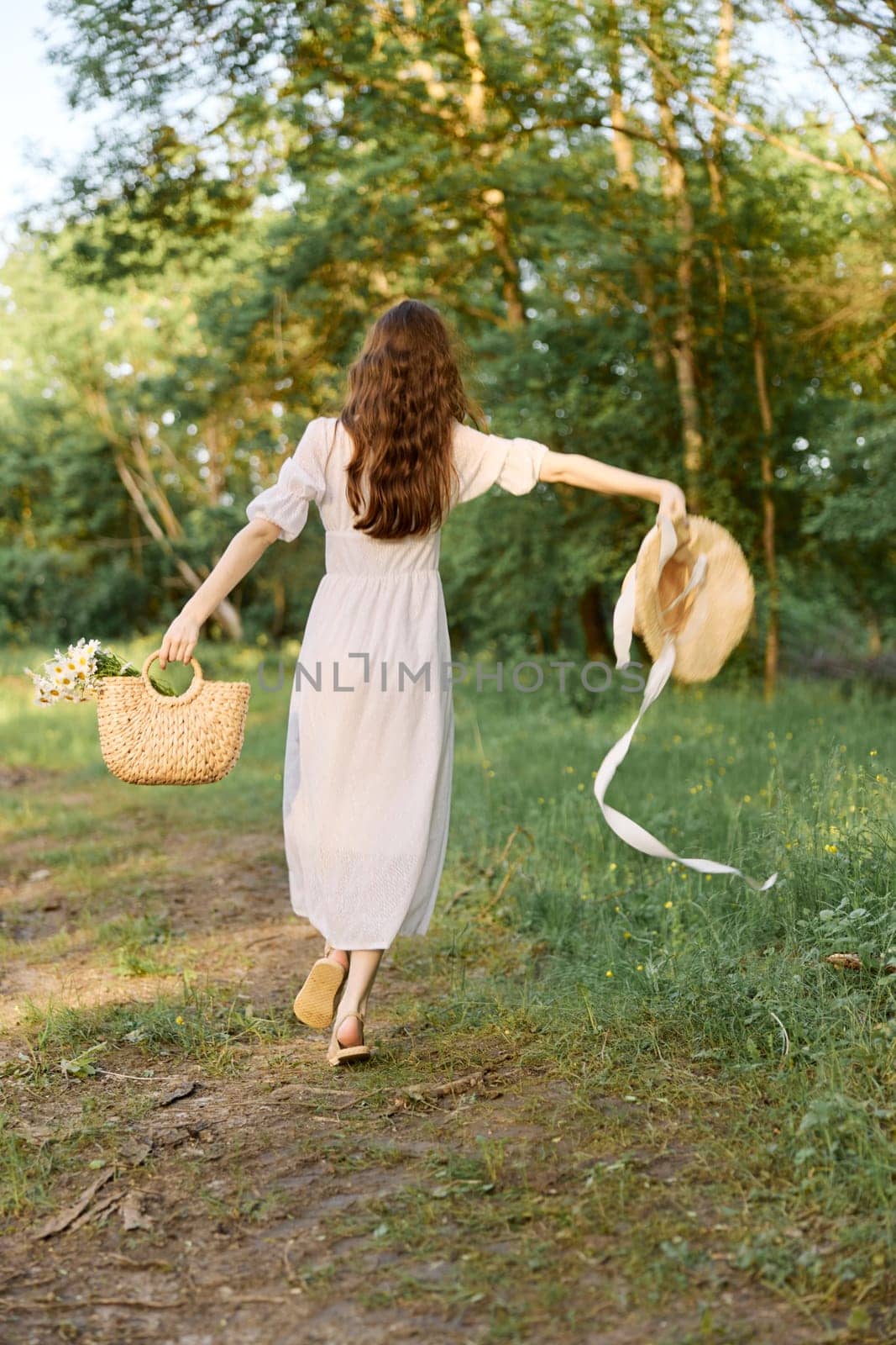 a woman in a long summer dress walks along a forest path holding a wicker hat with flowing ribbons in her hands. High quality photo
