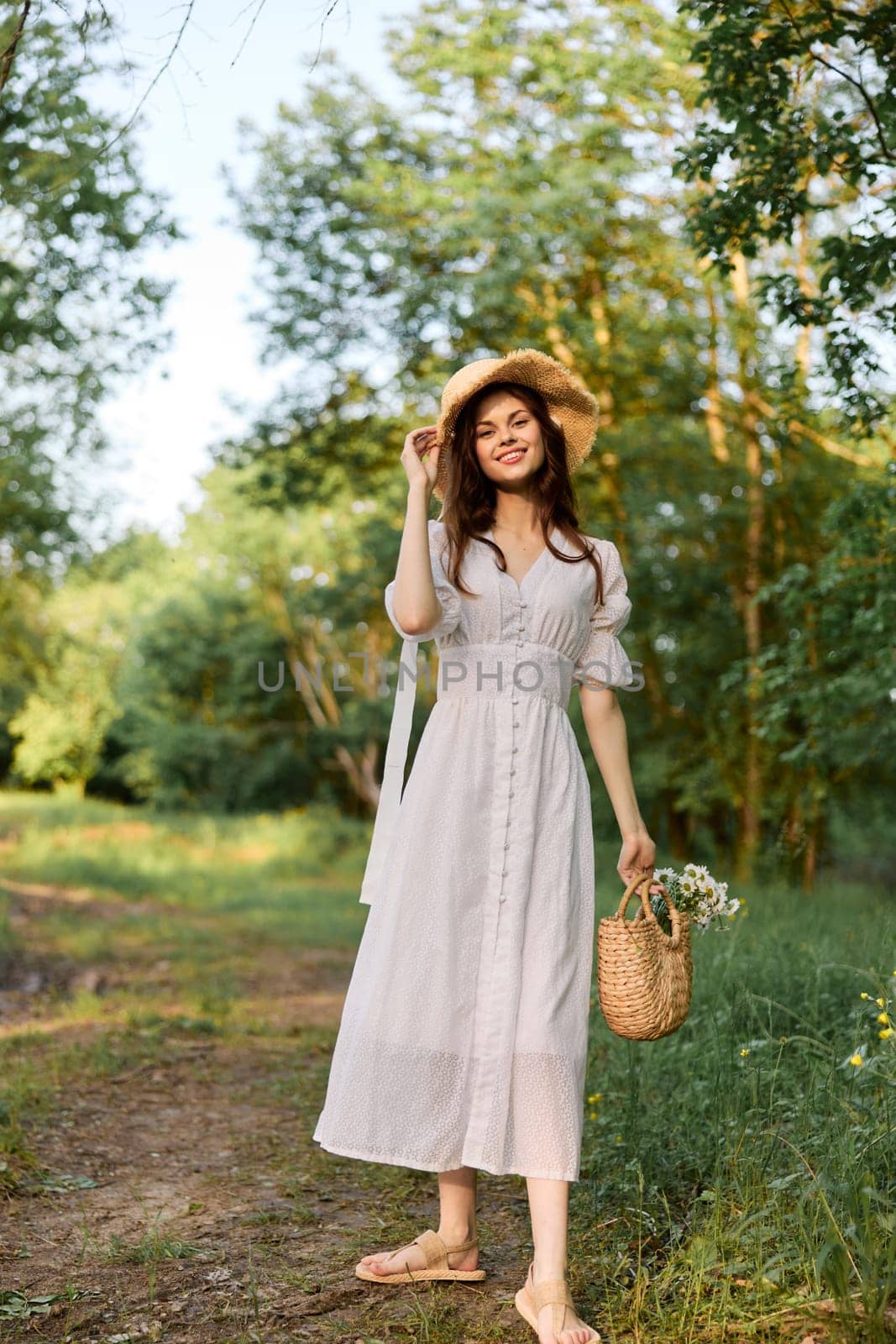 a woman in a light summer dress and a wicker hat stands in the forest with a basket of daisies in her hands. High quality photo