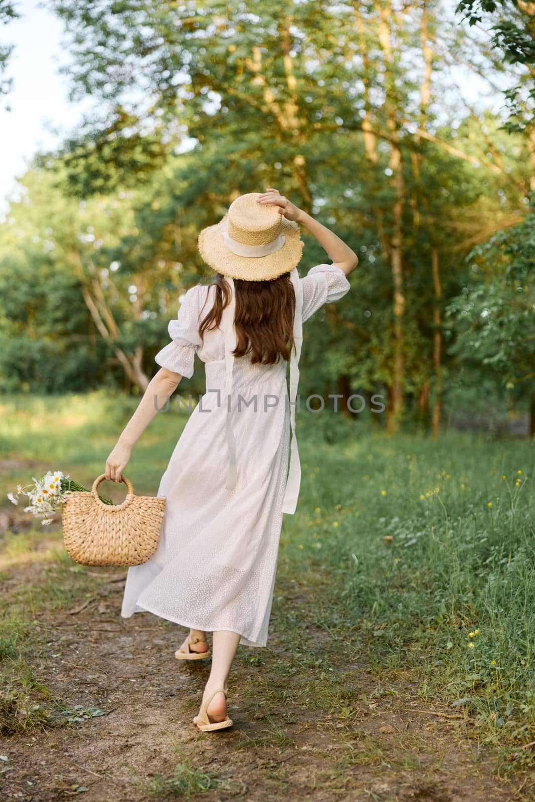 a woman in a long summer dress walks along a forest path holding a wicker hat on her head with her hand. High quality photo